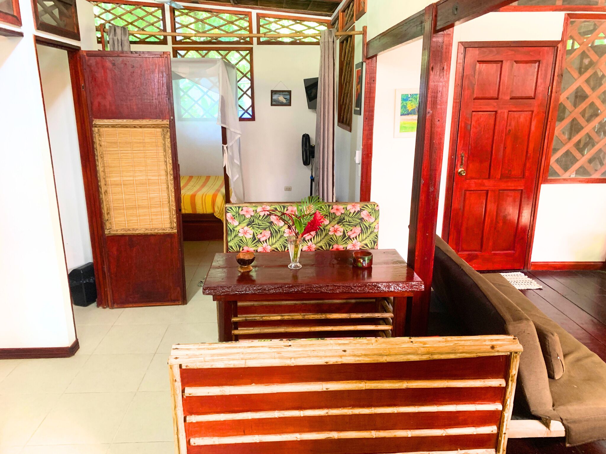 A room with a wooden bench, a table with a floral chair, a bed with a striped bedspread, and a red door.
