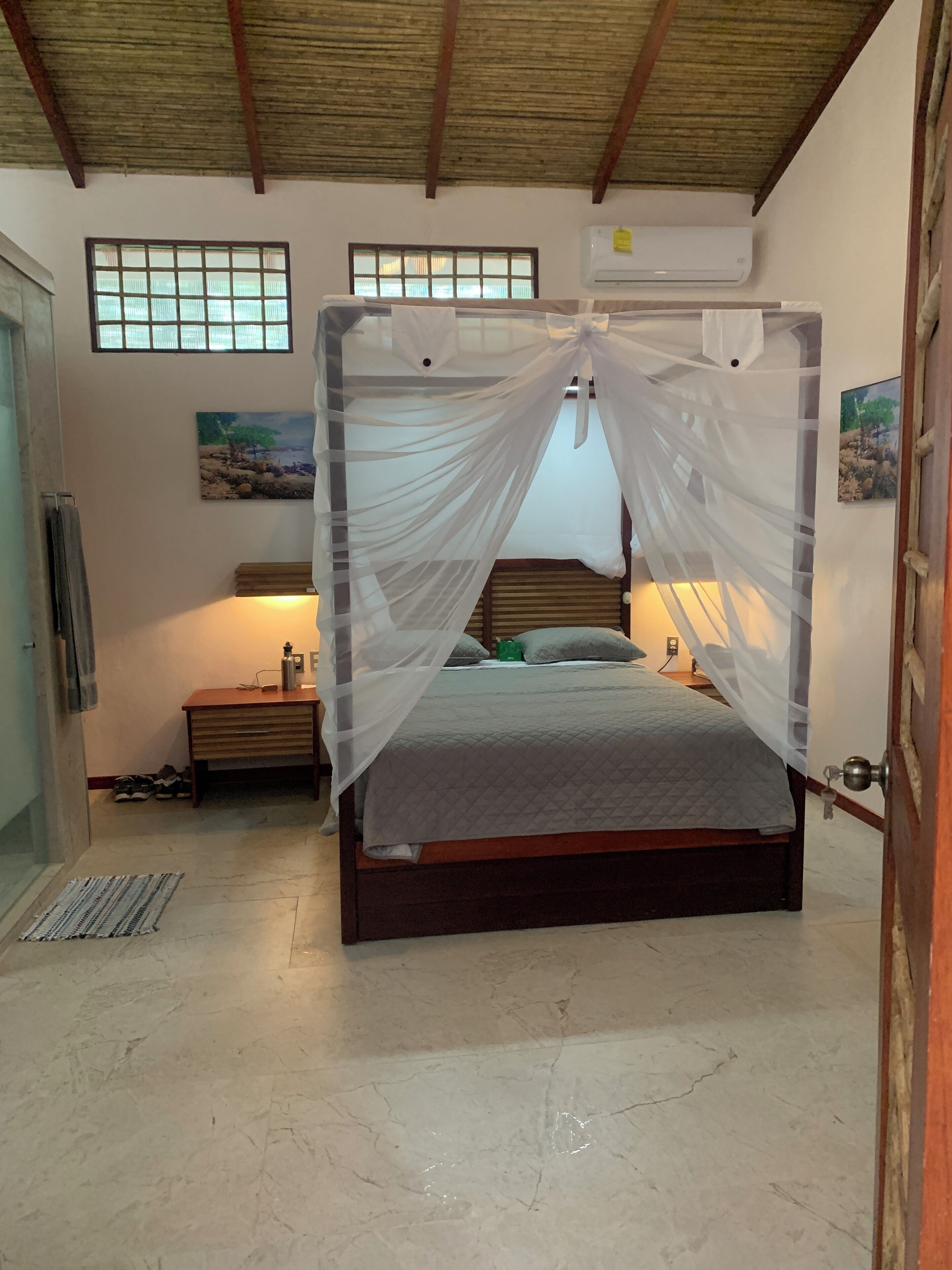 A bedroom with a bed under a mosquito net, a nightstand with a lamp, and a painting on the wall.