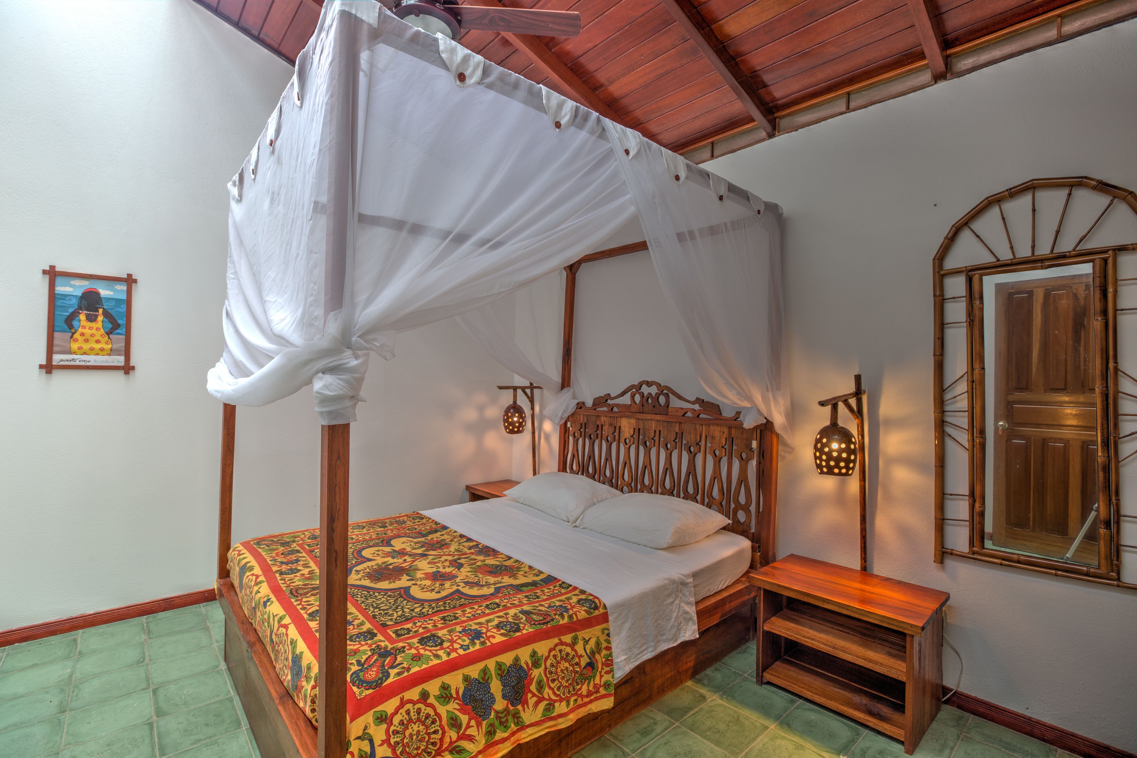 A four-poster bed with a canopy, a wooden nightstand, a wall-mounted lamp, and a framed picture on the wall.