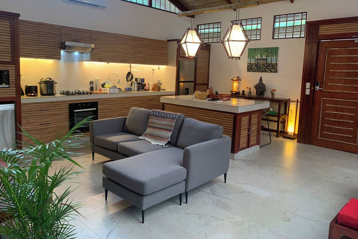 A modern kitchen with a central island, a dining area with a sofa, and a large window providing natural light.