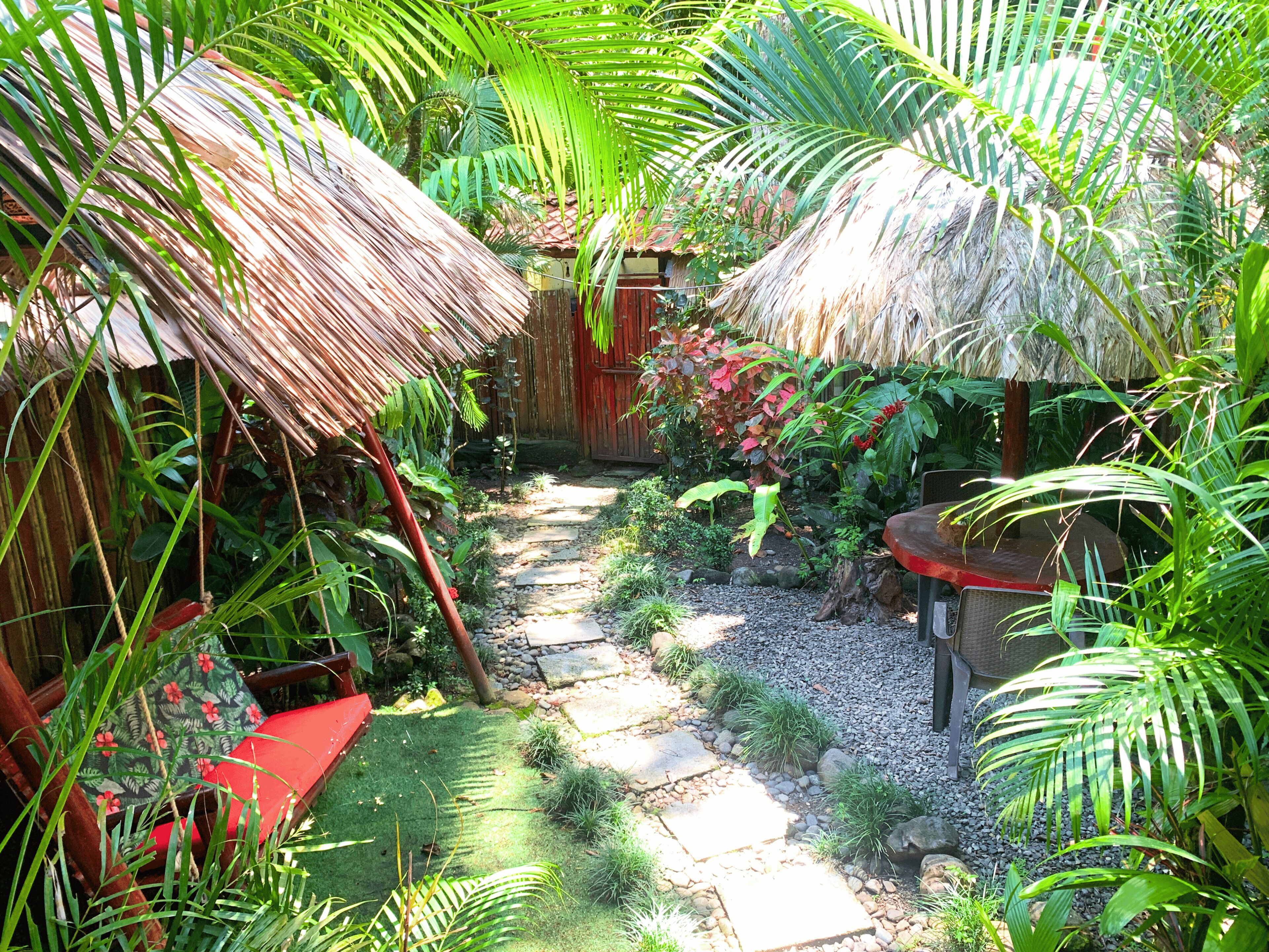 Ein tropischer Garten mit strohgedeckten Bauten, einem Kiesweg und üppiger Vegetation.