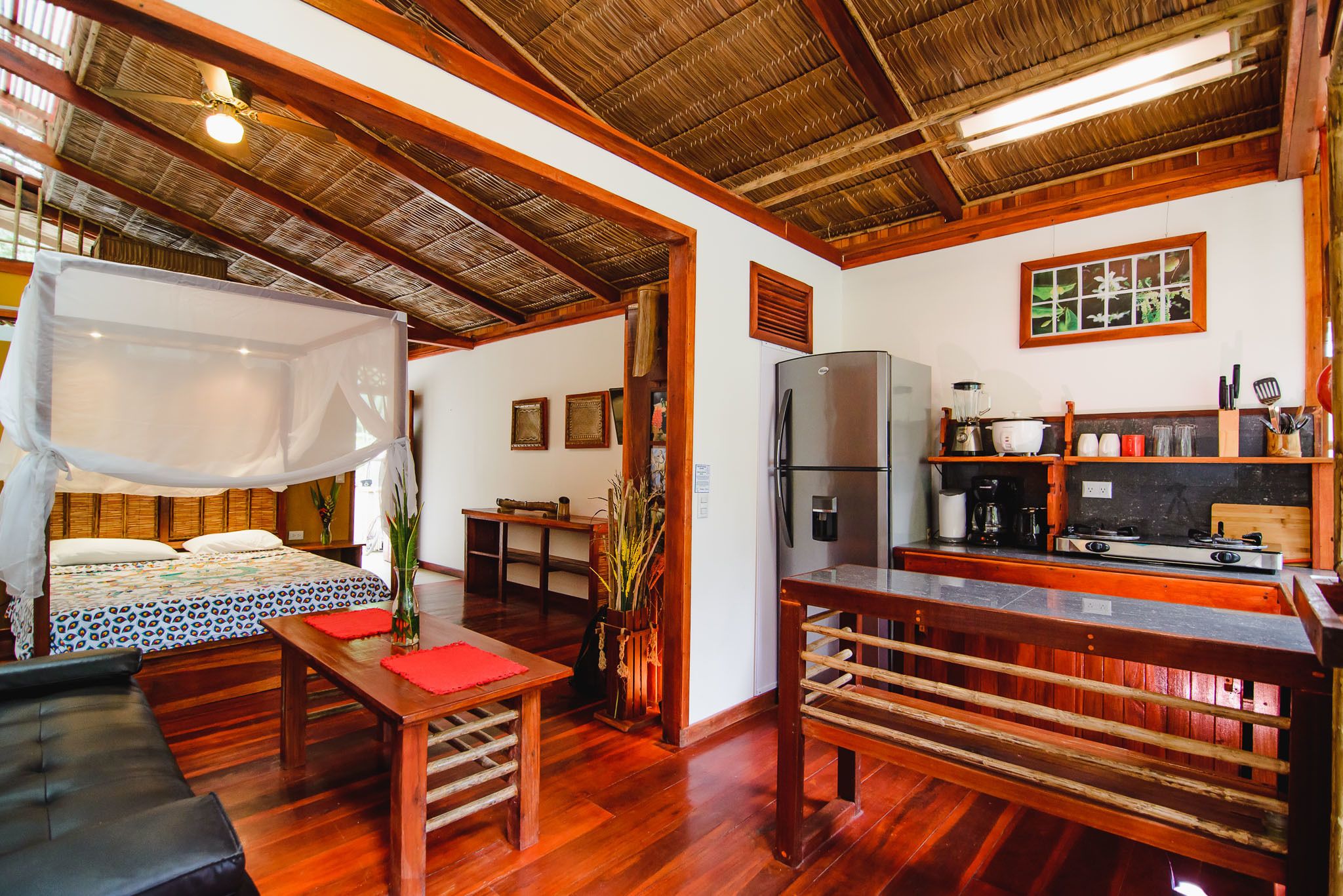 A room with a bed, a wooden table, a bench, a kitchen area with a refrigerator and stove, and a ceiling fan.