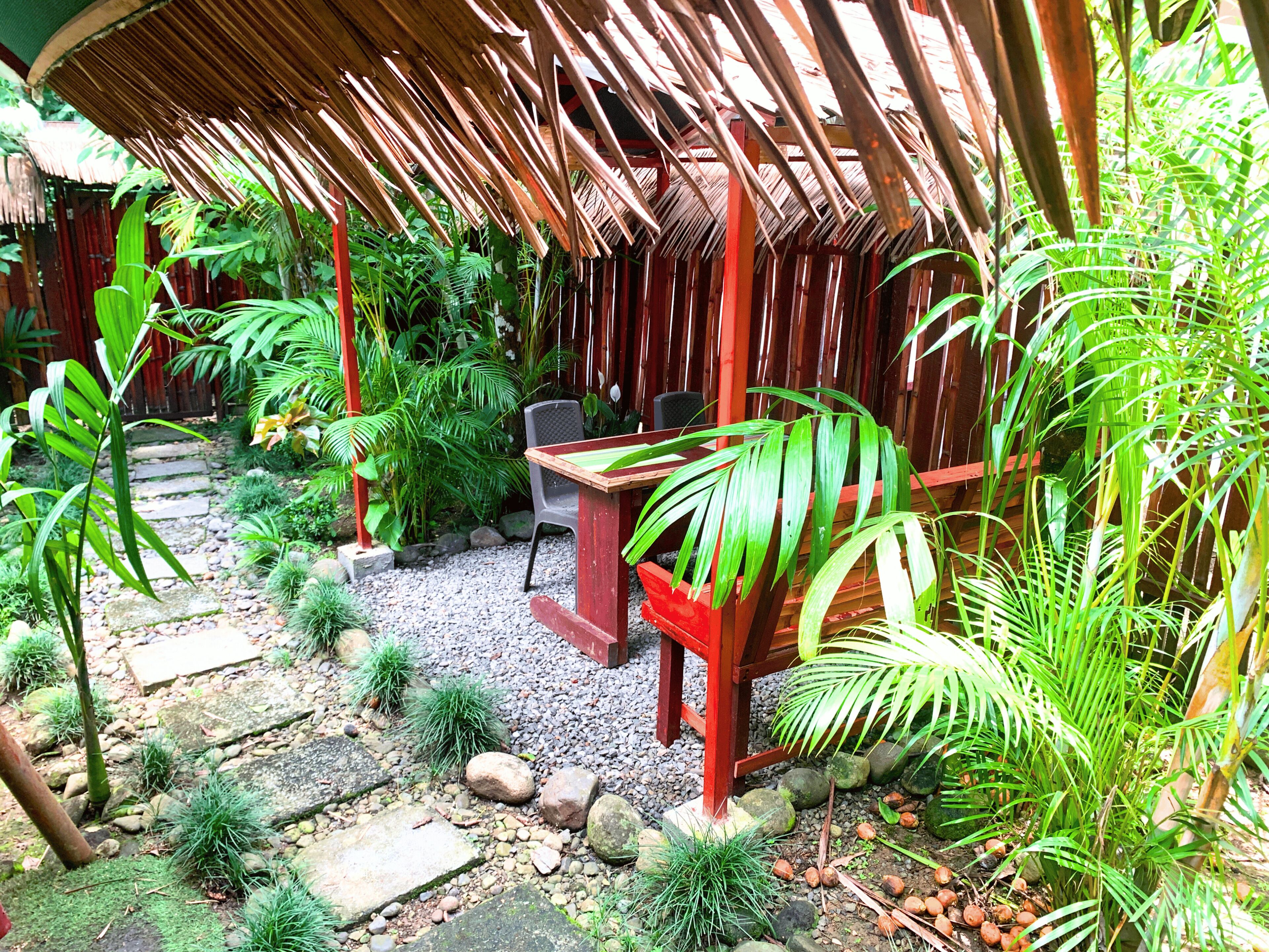 Un espace extérieur aménagé, abrité et doté d’une table et de chaises, entouré d’une végétation luxuriante et bordé d’un sentier en pierre.
