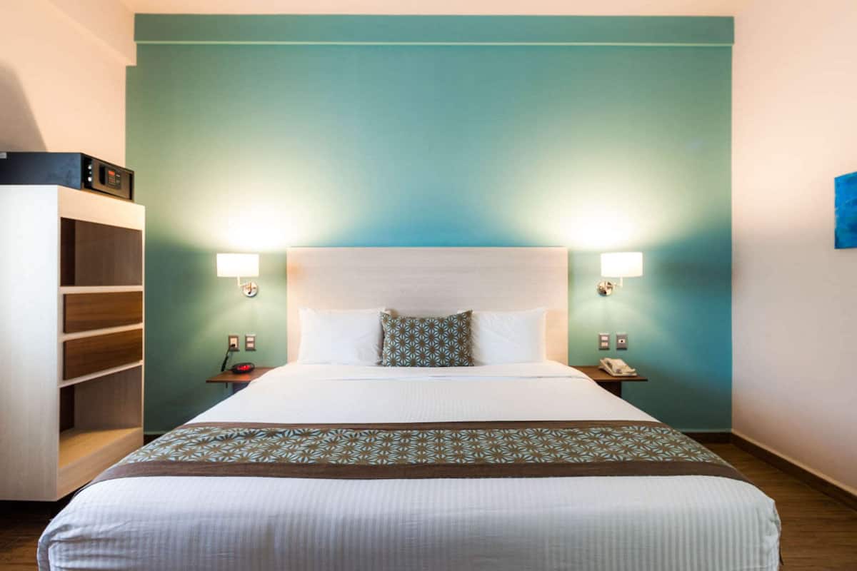 A hotel room with a bed, two bedside lamps, a nightstand, a telephone, and a shelf with a box on it.