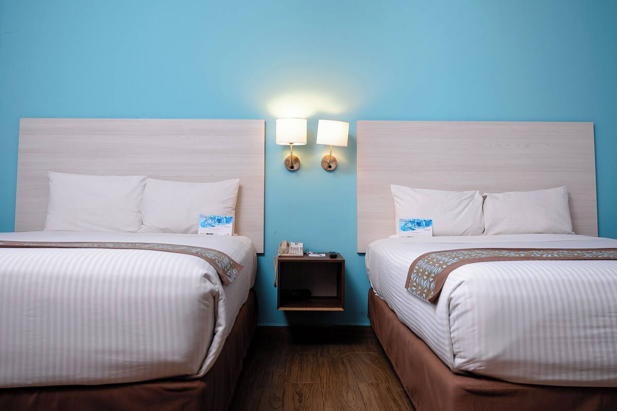 A hotel room with two beds, a nightstand with a lamp, and a blue wall.