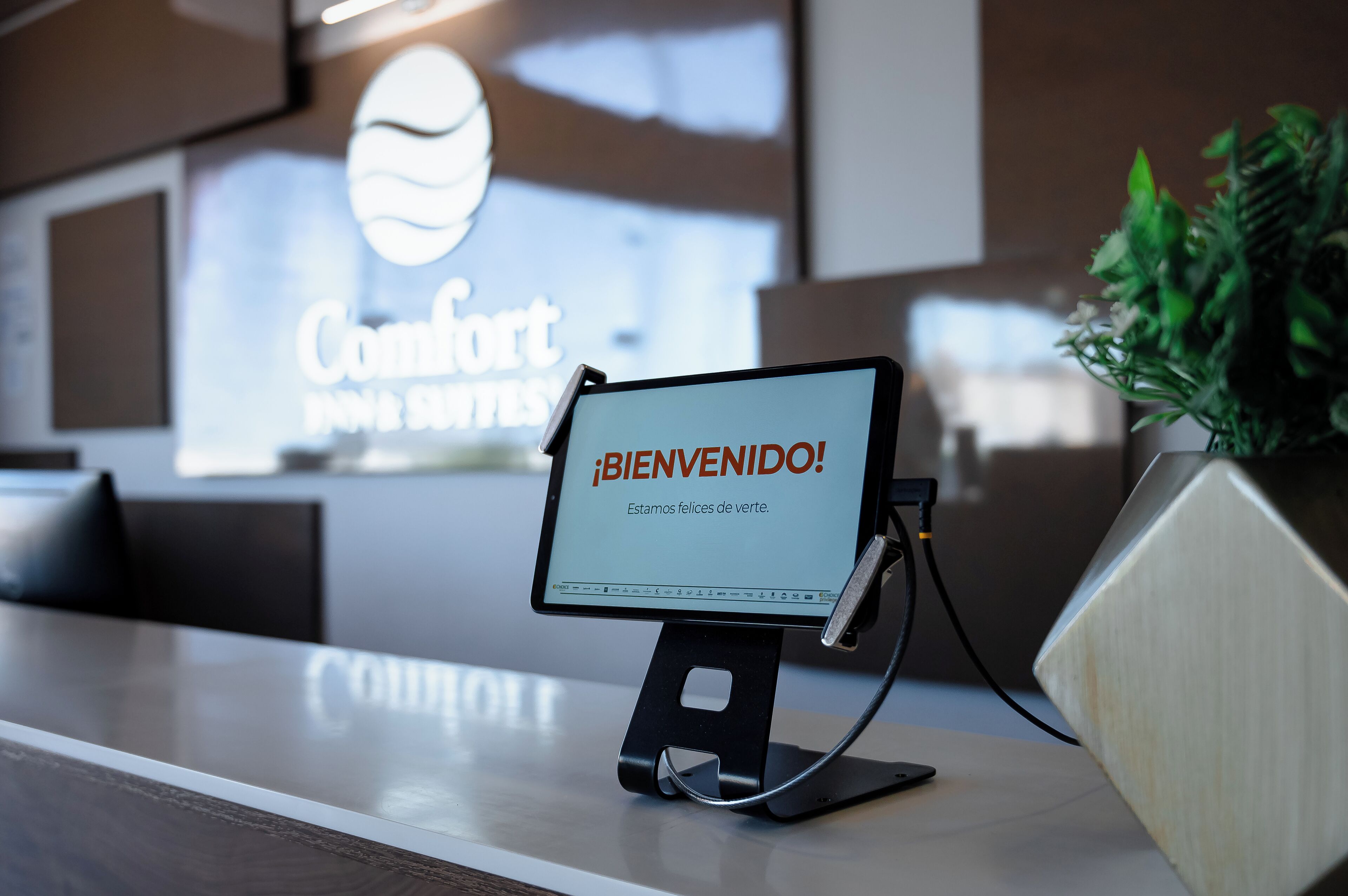 En receptionsdisk med en digital velkomstskærm, der viser "¡BIENVENIDO! Estamos felices de verte." og et skilt i baggrunden med teksten "Comfort for your senses.".