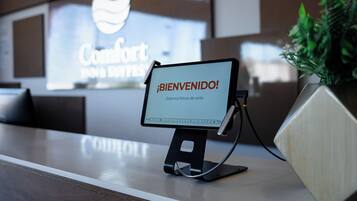 Uma mesa de recepção com uma tela digital exibindo "¡BIENVENIDO! Estamos felizes em vê-lo." e uma placa ao fundo com a inscrição "Conforto para seus sentidos".
