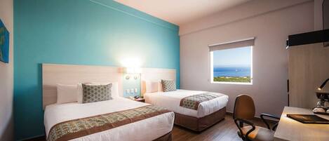 A hotel room with two beds, a desk, and a view of the sea.