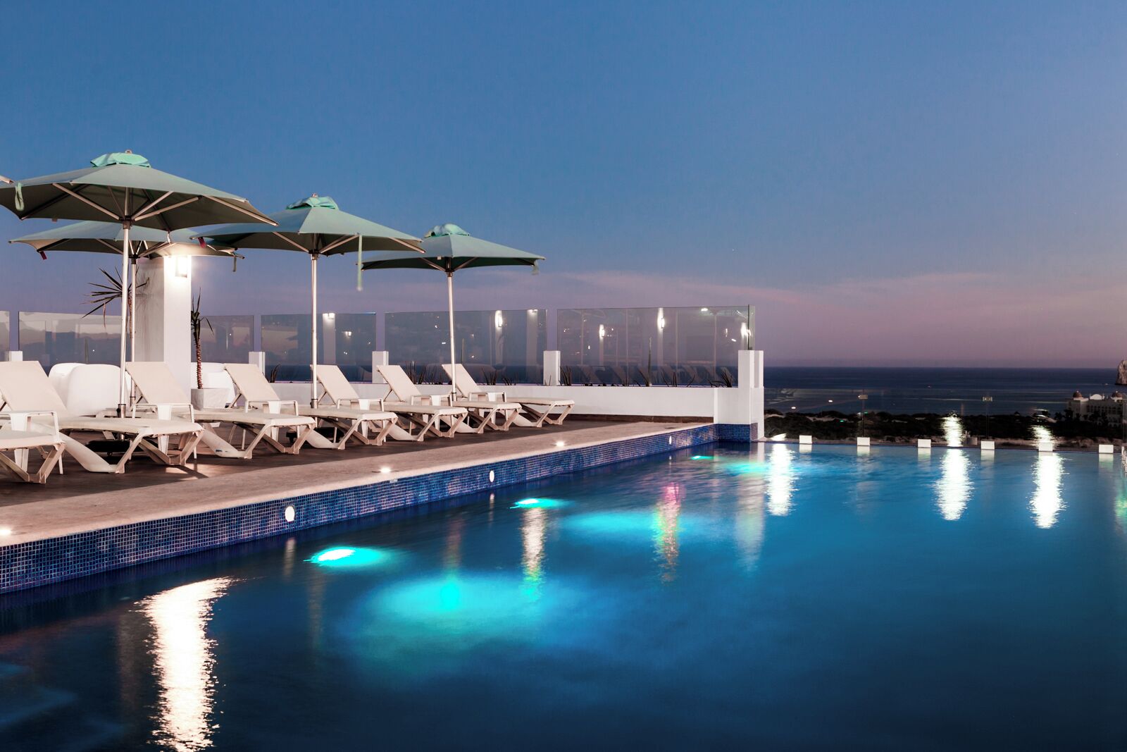 Un área junto a la piscina con sillones de descanso y sombrillas, vista despejada al océano y una piscina con baldosas.