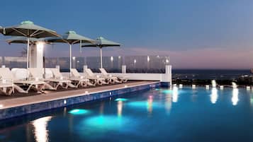 Un área junto a la piscina con sillones de descanso y sombrillas, vista despejada al océano y una piscina con baldosas.