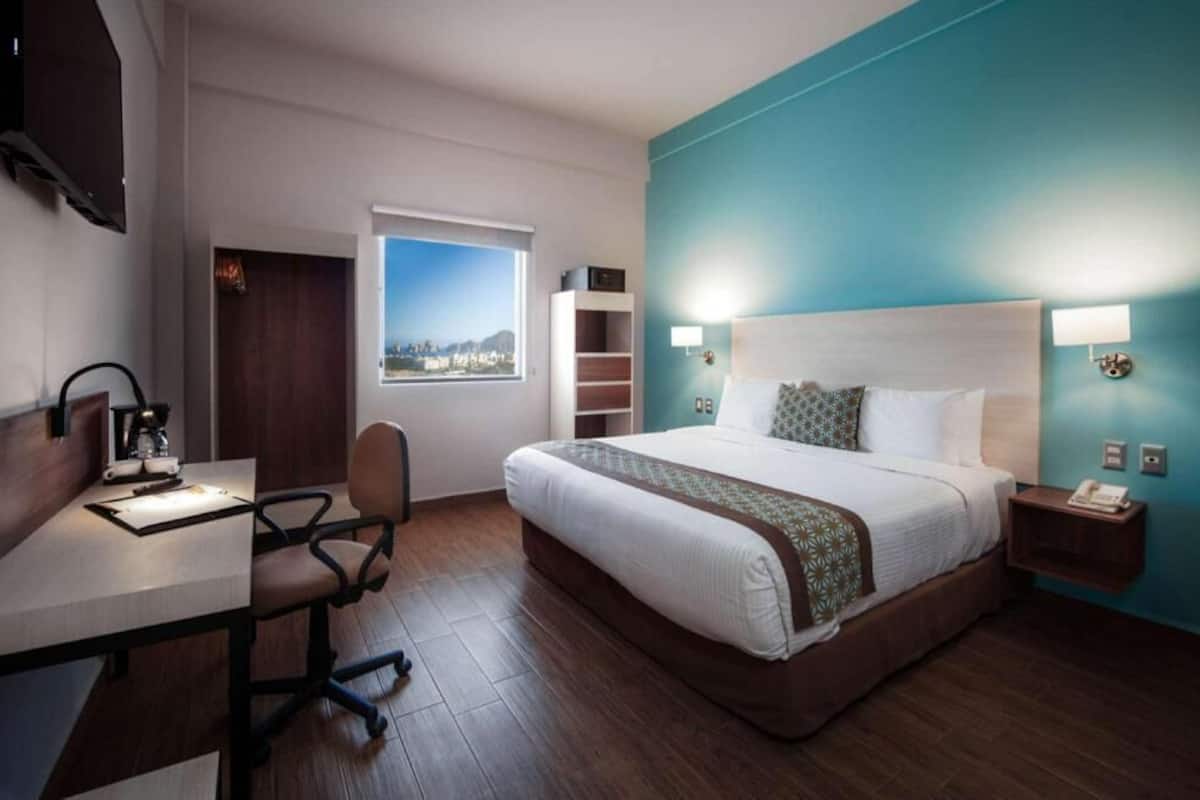 A hotel room with a bed, desk, chair, and a view of the city through a window.