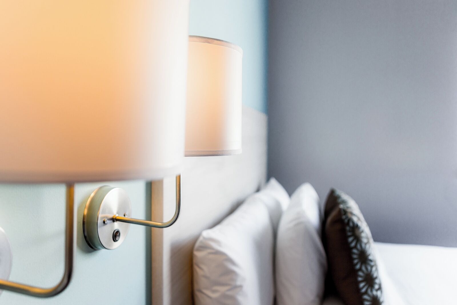 Een close-up van een wandlamp met een witte kap en een metalen voet, geplaatst naast een bed met witte kussens en een kussen met een donker patroon.