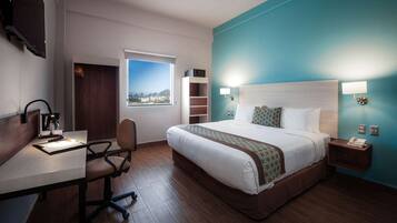 Quarto de hotel com uma cama grande, uma escrivaninha com cadeira, televisão, janela com vista para a cidade e uma parede azul como detalhe.