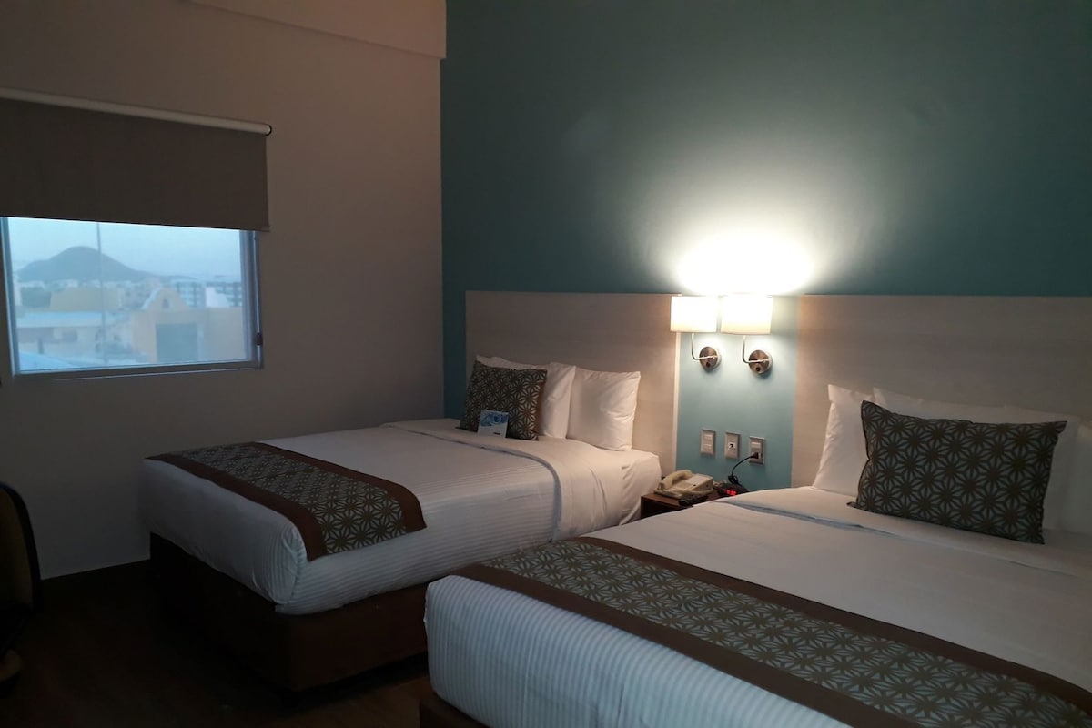 A hotel room with two beds, a window, a nightstand, and a wall-mounted lamp.