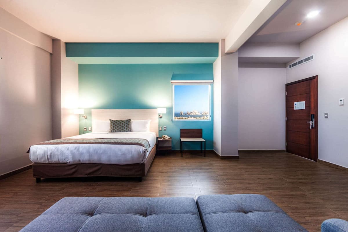 A modern hotel room with a large bed, a seating area, and a view of the city through a window.