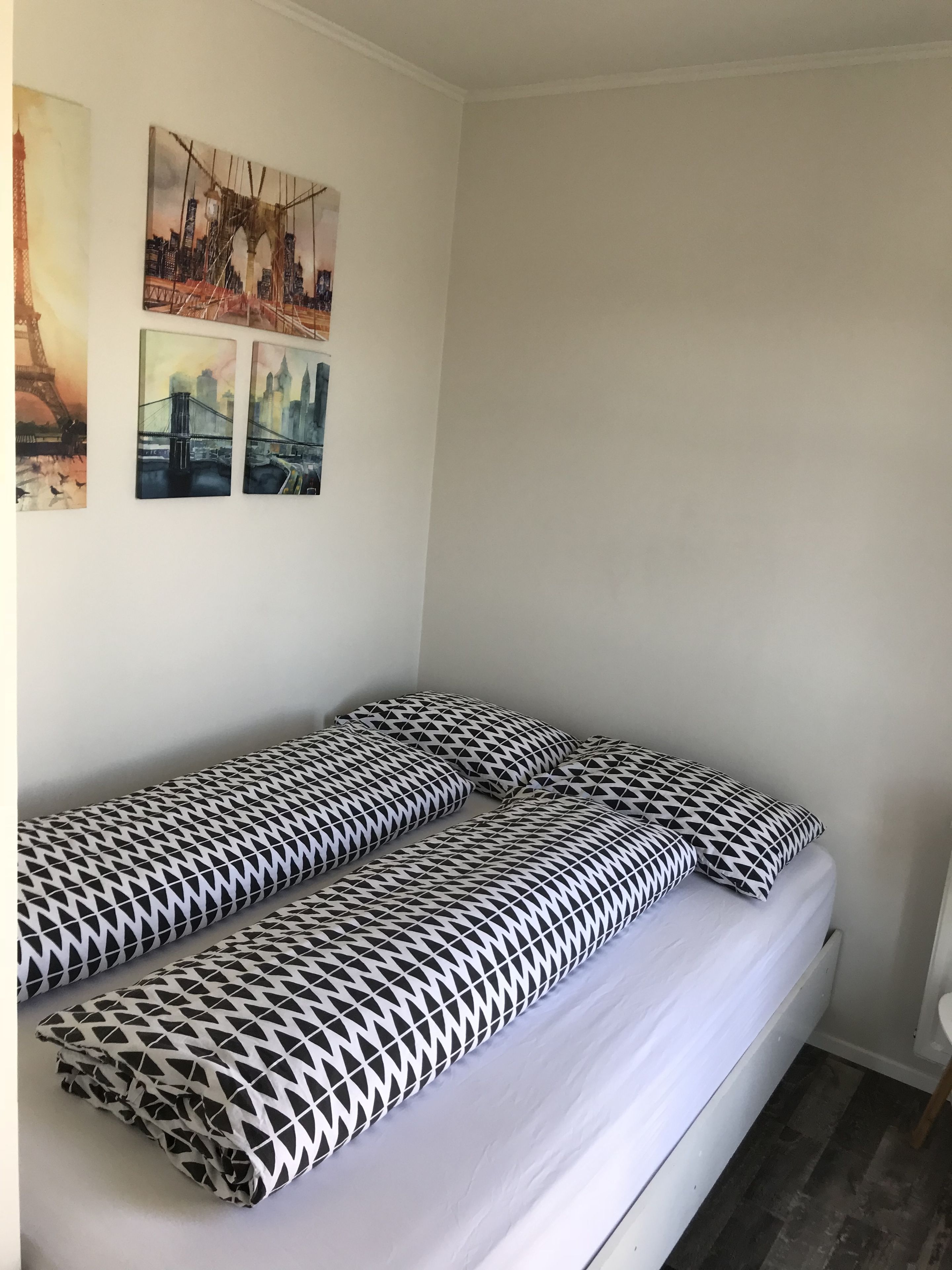 A single bed with patterned pillows, a white wall, and three cityscape paintings.