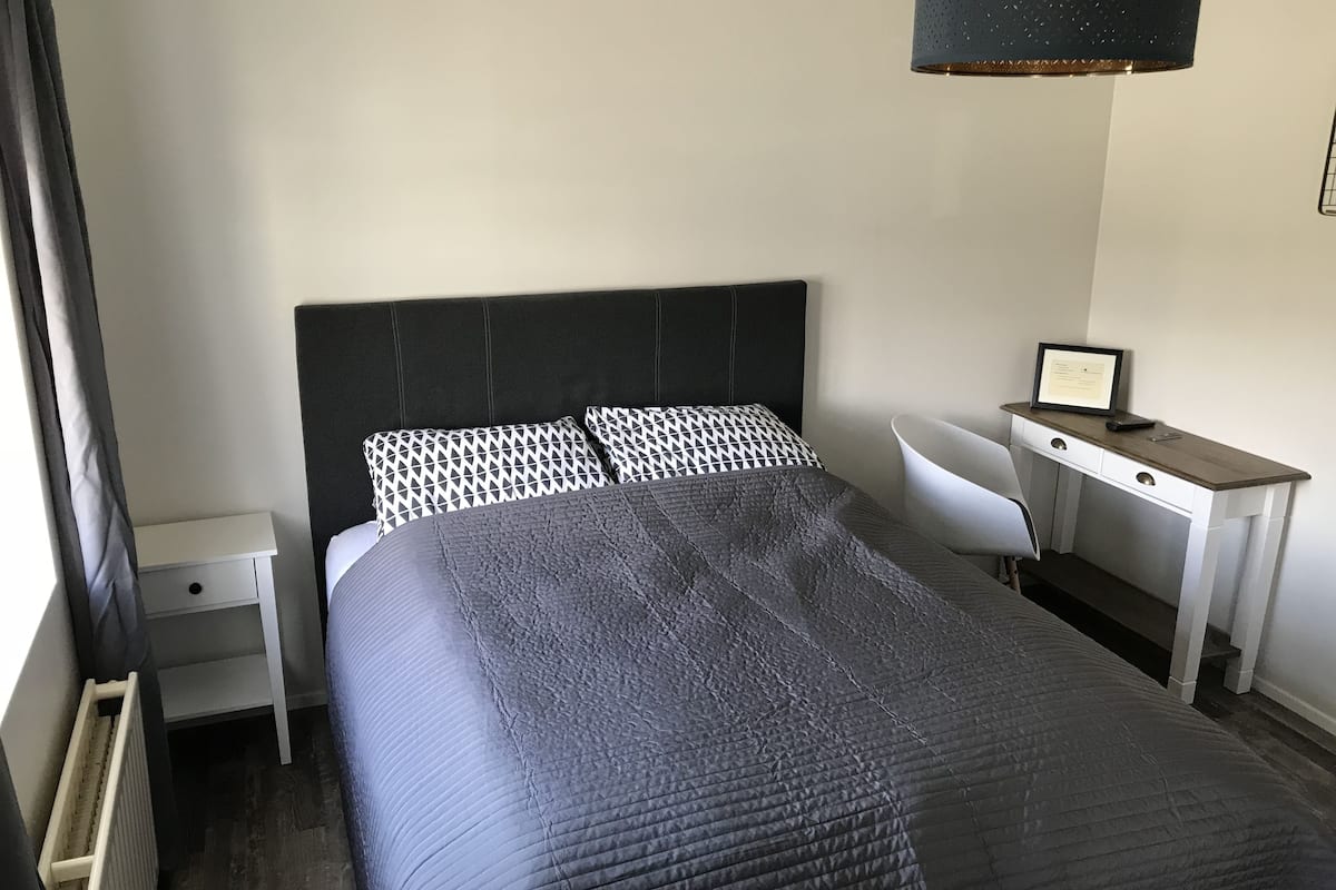 Una cama bien hecha con un cabecero oscuro, una mesita de noche con una lámpara y un escritorio con una silla.