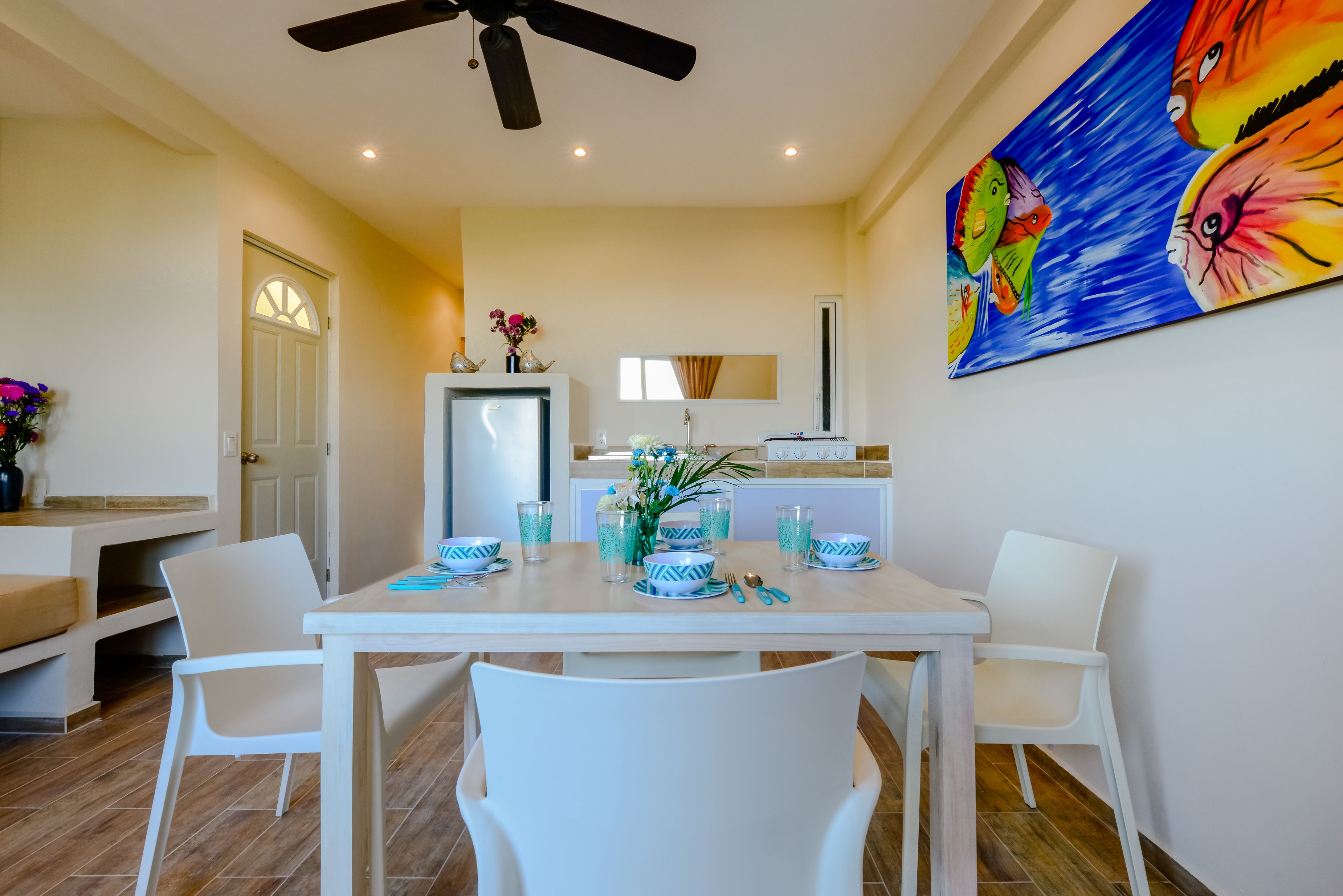 A dining area with a white table set for four, a ceiling fan, and a colorful painting on the wall.