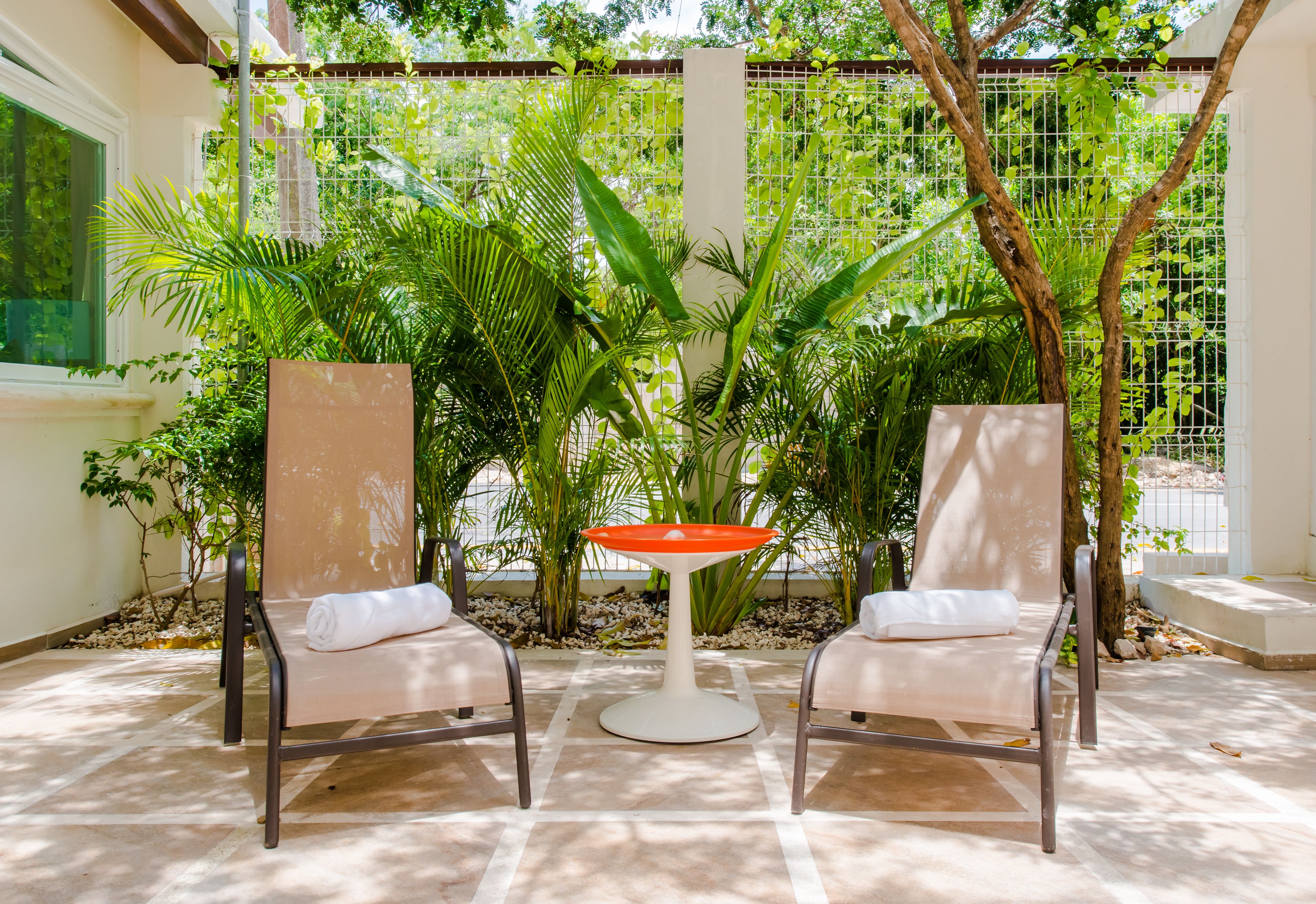 Two lounge chairs with white cushions, a round table with an orange top, and a backdrop of lush green plants and a white fence.
