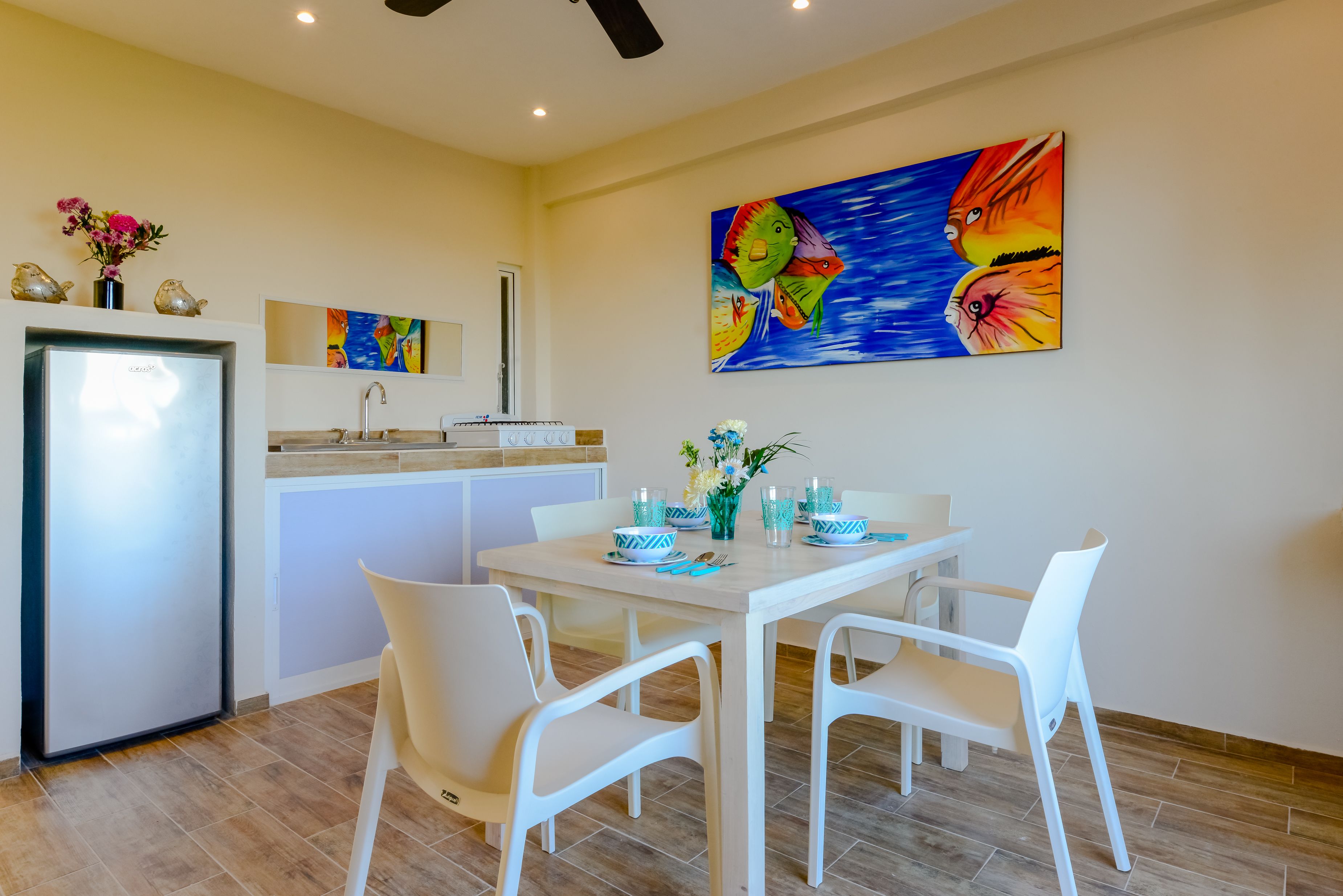 Una cocina moderna con refrigerador, lavamanos y comedor con sillas blancas y una mesa preparada para dos.
