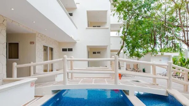 Una piscina con plataforma y barandillas blancas, rodeada por un edificio blanco con balcones y un árbol.