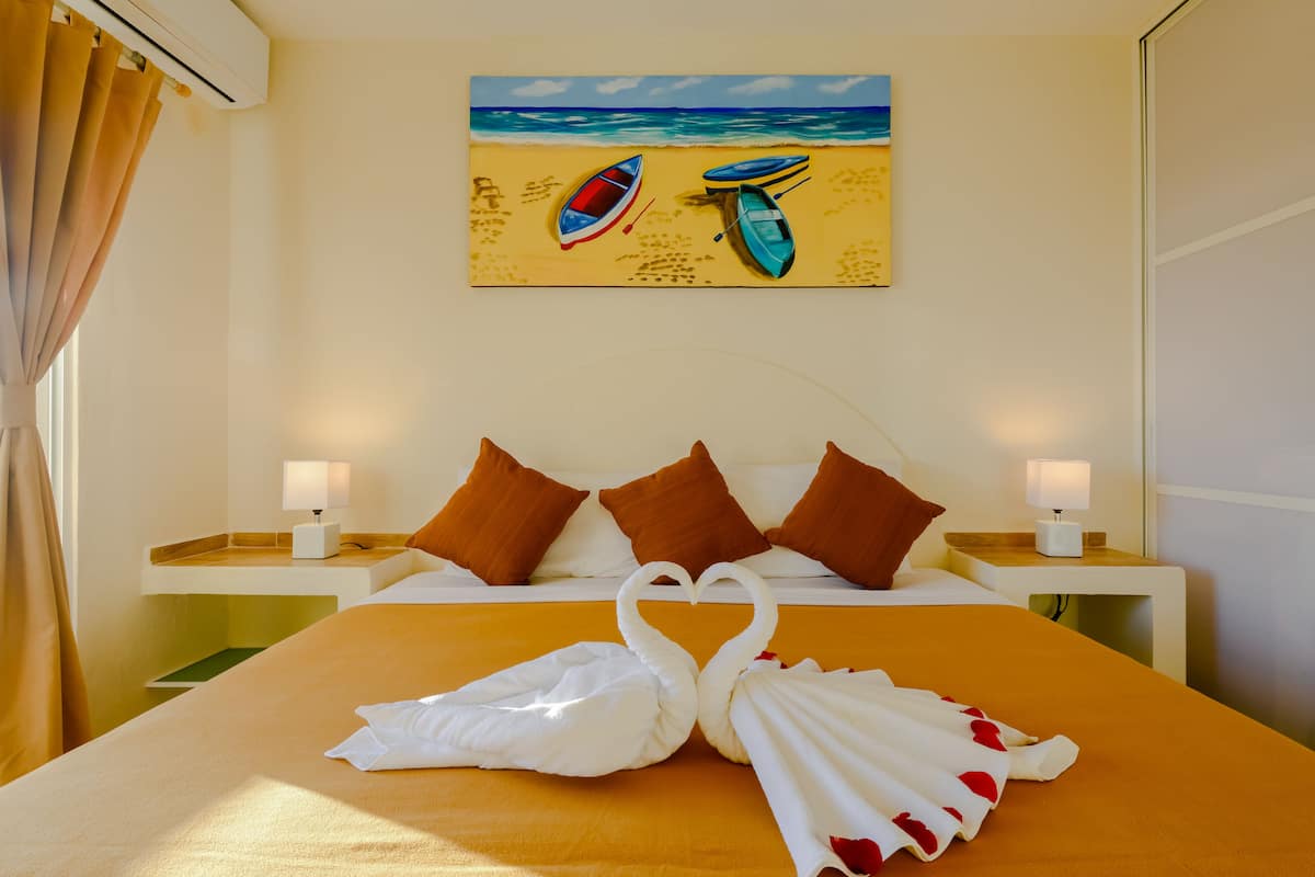 Una habitación de hotel con cama, dos mesitas de noche, un cuadro de barcos en la playa y una colcha con forma de cisne.
