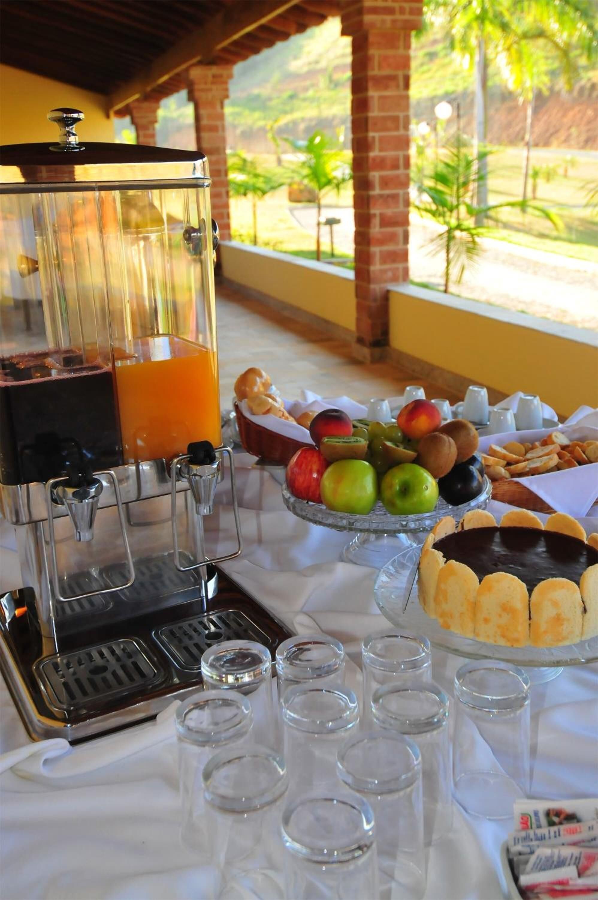 Buffet de café da manhã com dispensador de suco, tigela de frutas, pães e pequenos copos.