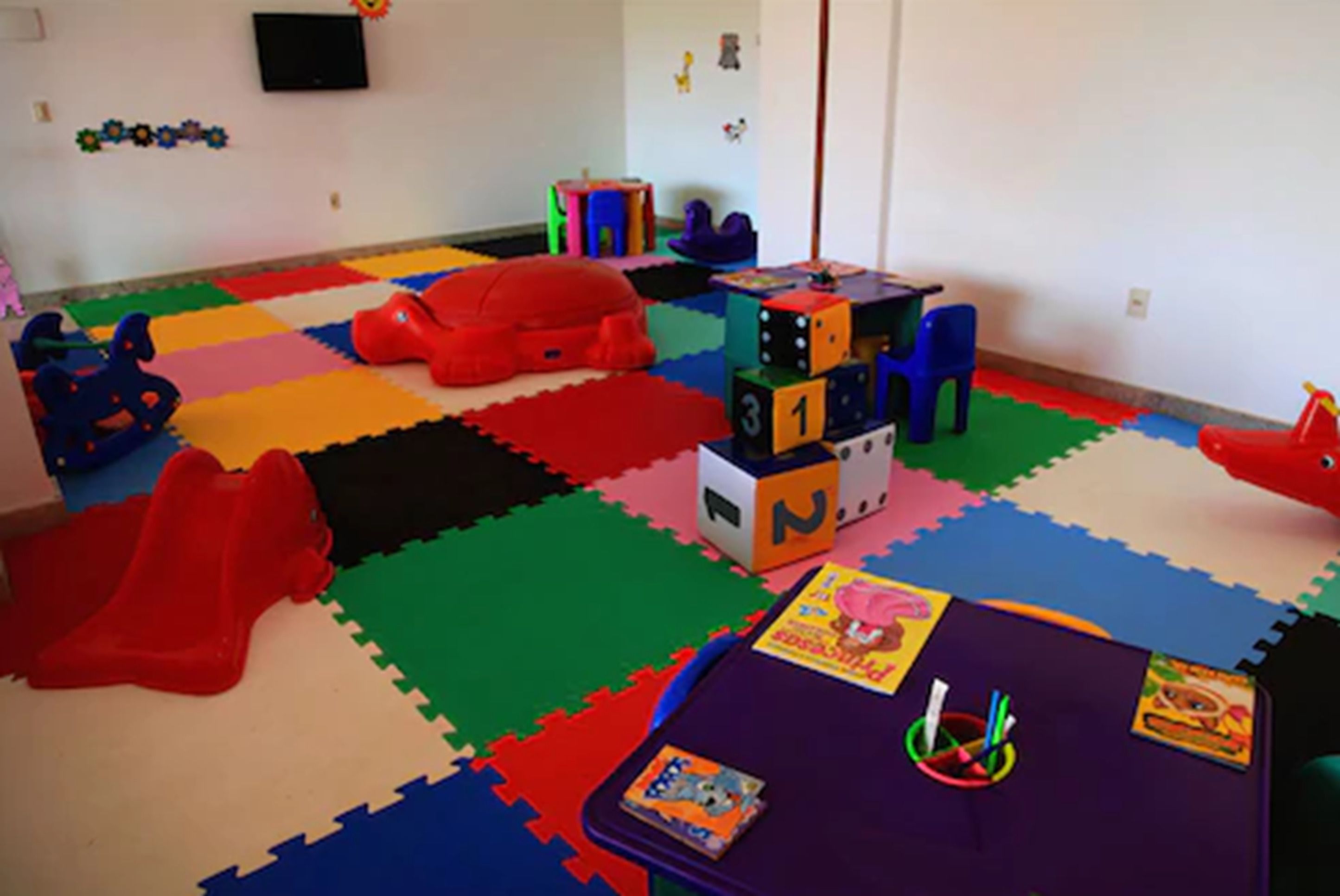 A playroom with colorful foam mats, a purple table, and various toys including blocks and a red car.