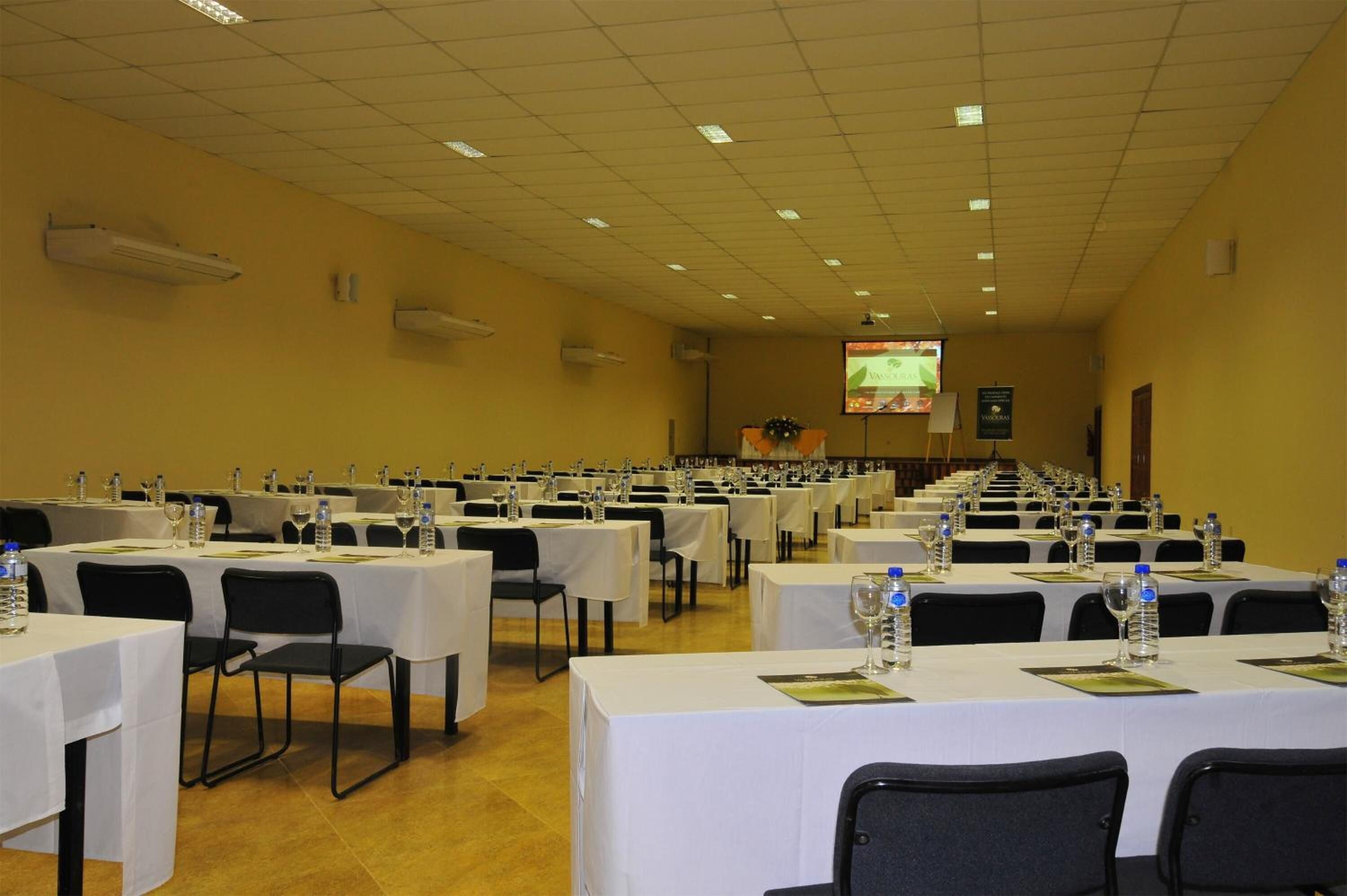 Sala de conferência com mesas dispostas em fileiras para uma reunião, equipada com garrafas de água e porta-nomes.