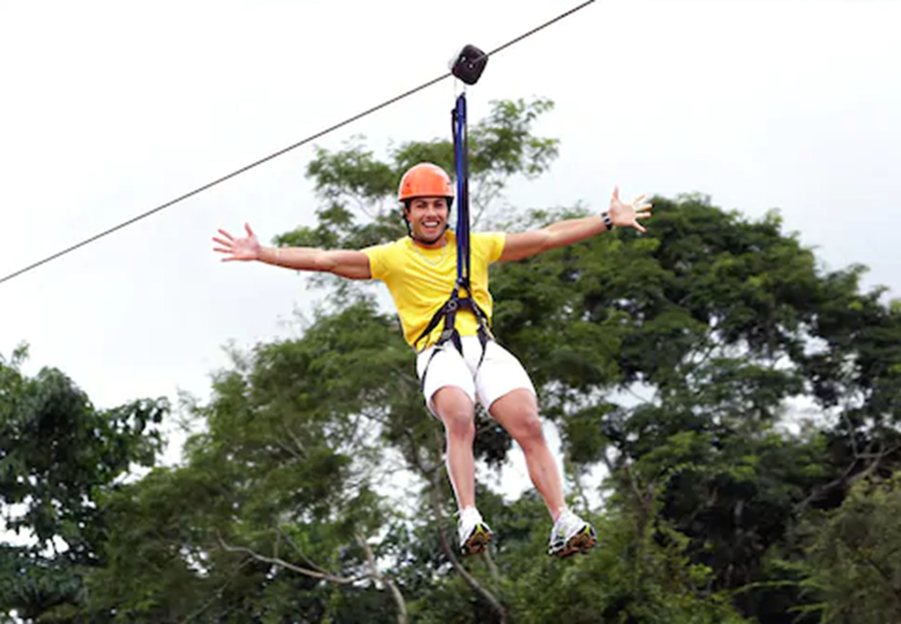 A person in a yellow shirt and white shorts suspended in mid-air, wearing a safety harness and helmet, likely engaged in an outdoor activity.