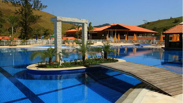 A swimming pool with a wooden deck, palm trees, and a building with a red roof in the background. - Vassouras Eco Resort (Vassouras)