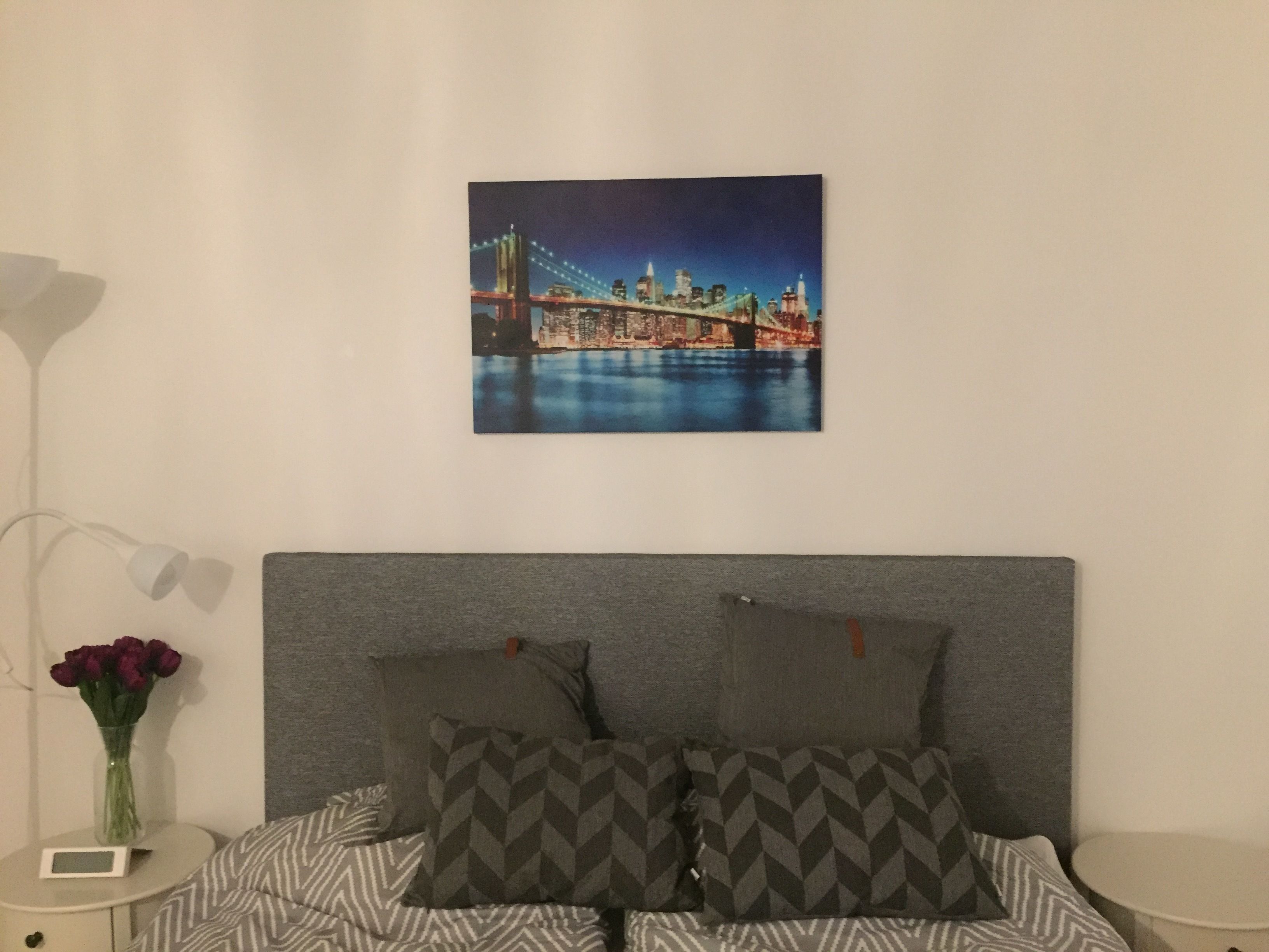 Un dormitorio con una cama, almohadas, una mesita de noche, una lámpara y un cuadro en la pared de un paisaje urbano.