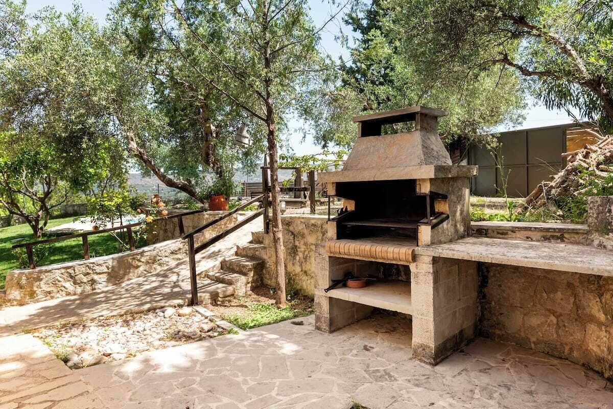 An outdoor stone barbecue area with steps leading up to it, surrounded by trees and a view of the sea.