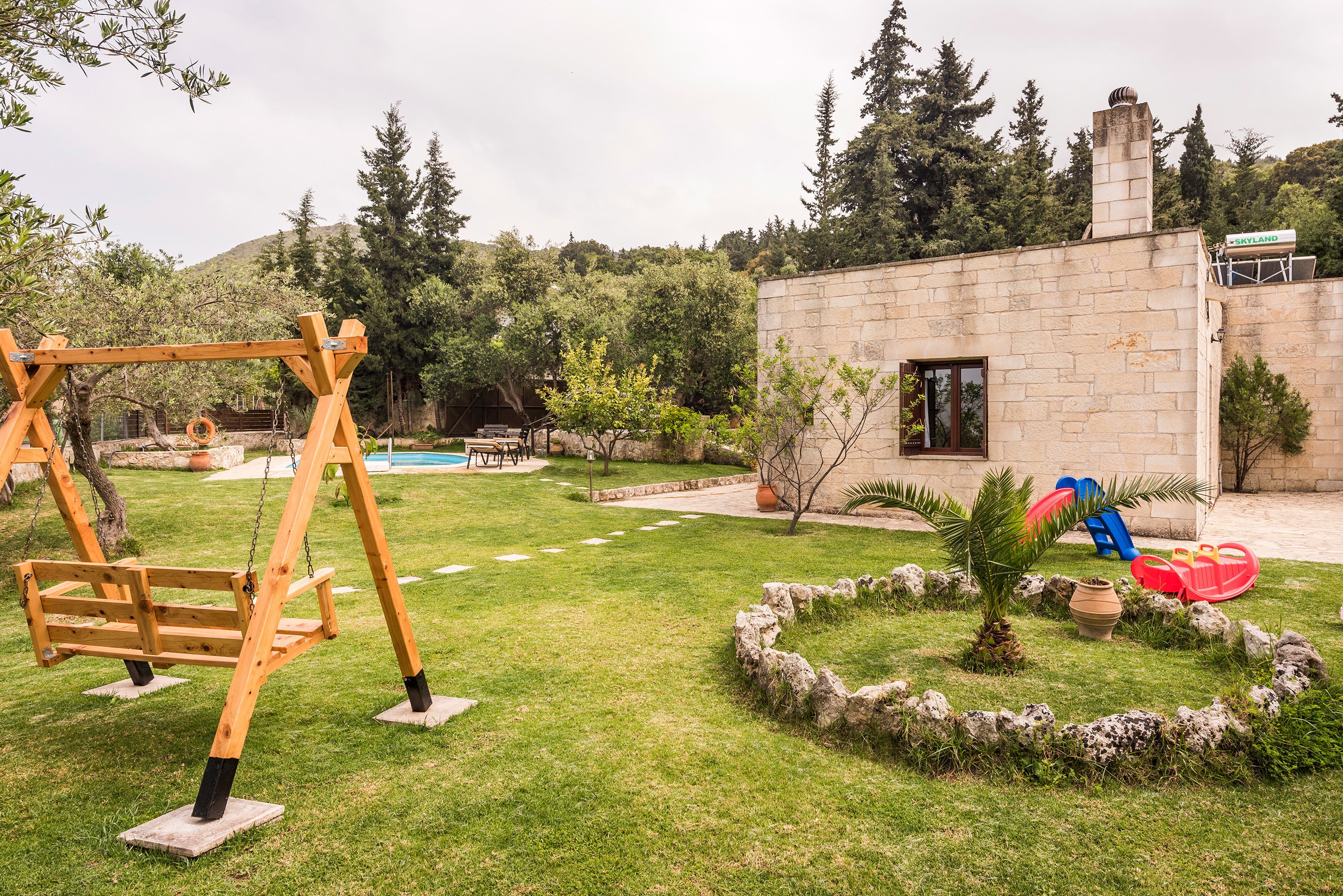 A backyard with a wooden swing set, a stone building, a small palm tree, and a rock garden.