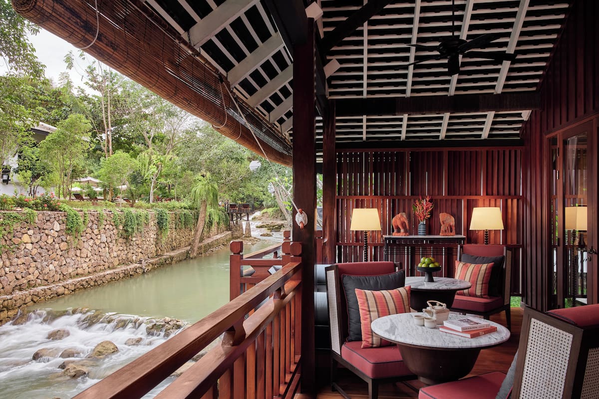 Une terrasse en bois offrant une vue sur une rivière, un mur en pierre et un espace détente avec des tables et des chaises.