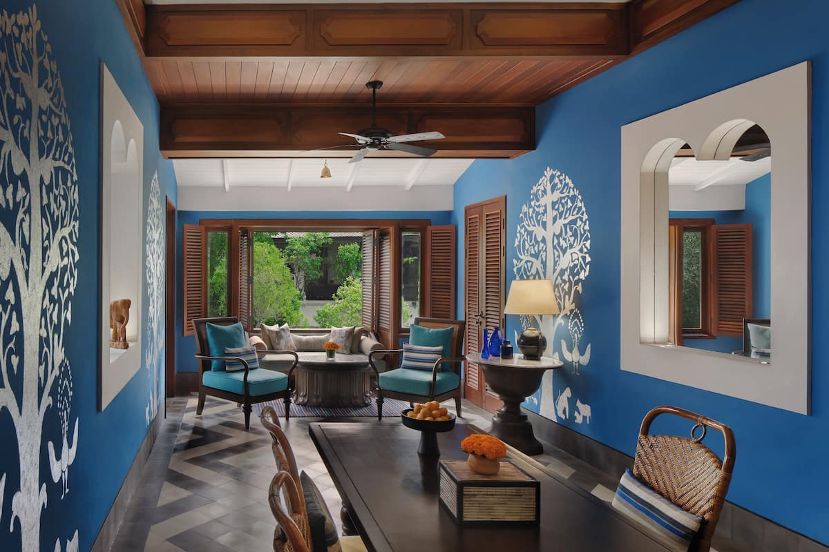 Une pièce avec un mur bleu, un plafond en bois et une vue sur la verdure à l’extérieur. On y trouve deux chaises, une table et une lampe.