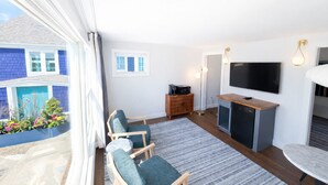 A modern living room with a TV, seating, and a view of a blue building. - The Foxberry - A Digital Concierge Guesthouse (Provincetown)