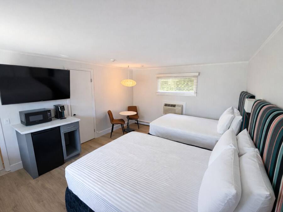 A hotel room with a bed, a desk with a microwave, a TV, a chair, a window with blinds, and a striped headboard.