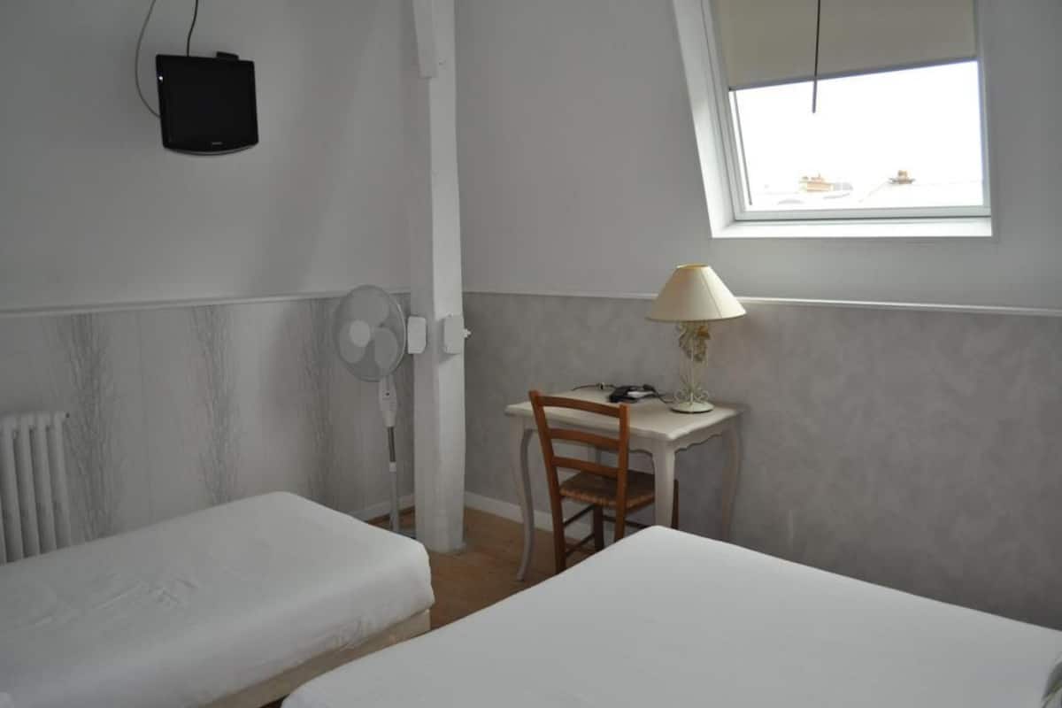 A small, simple room with two single beds, a desk with a chair, a lamp, a wall-mounted fan, and a window with a view.