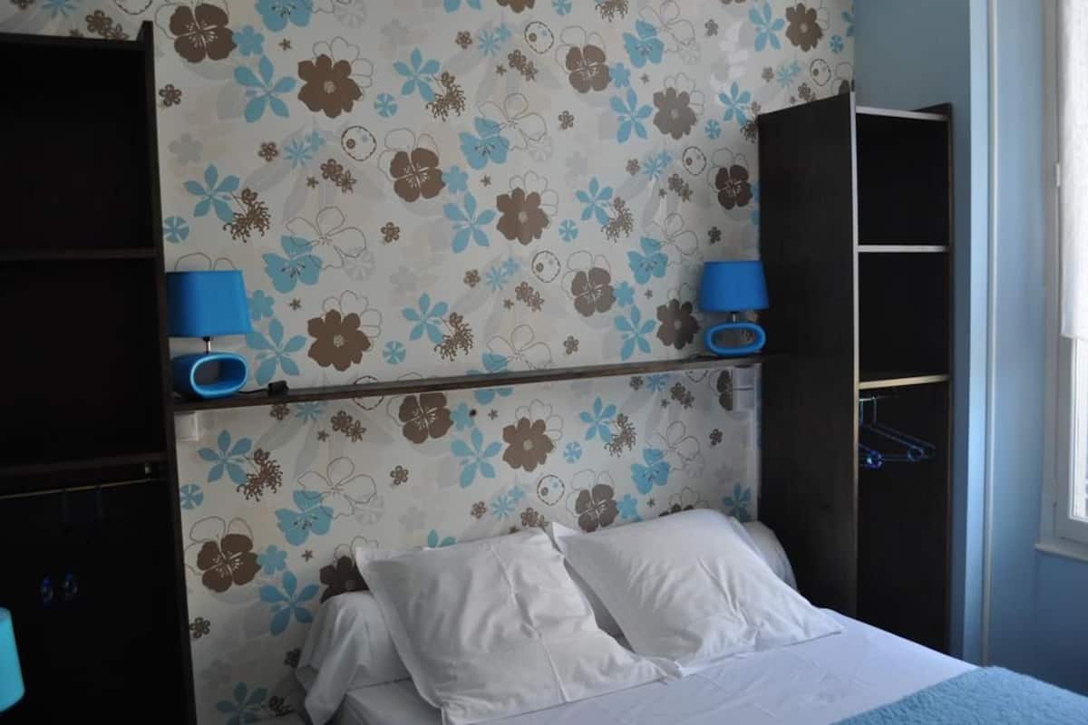 A bedroom with a bed, white pillows, a blue bedside lamp, a wardrobe, and a floral-patterned wall.