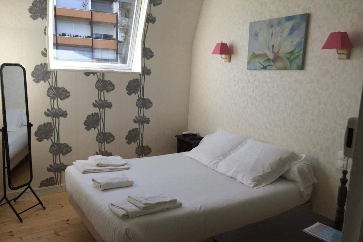A hotel room with a bed, a bedside table, a nightstand, a mirror, a window, and a painting on the wall.