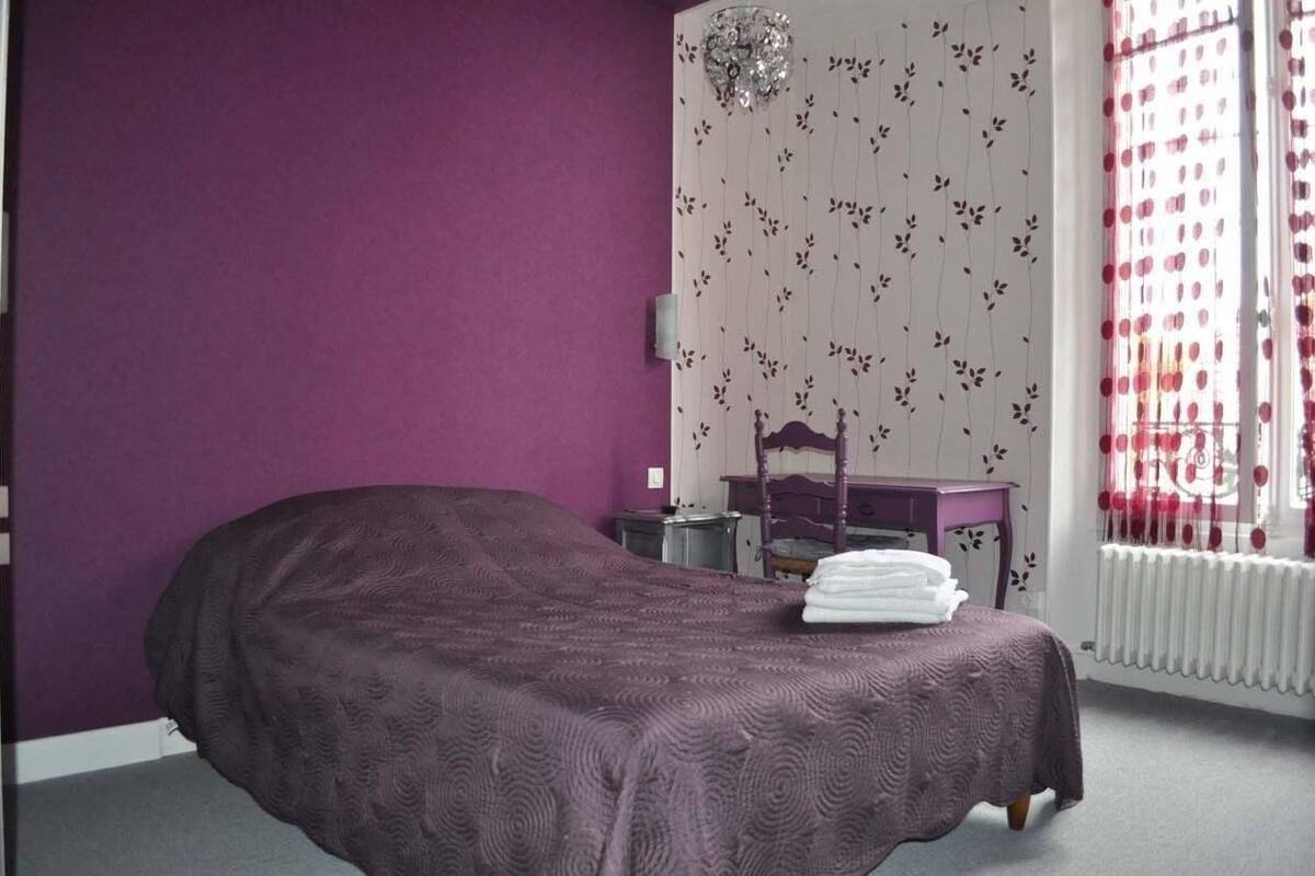 A bedroom with a purple wall, a bed with a purple bedspread, a wooden nightstand, a dresser, and a window with red curtains.