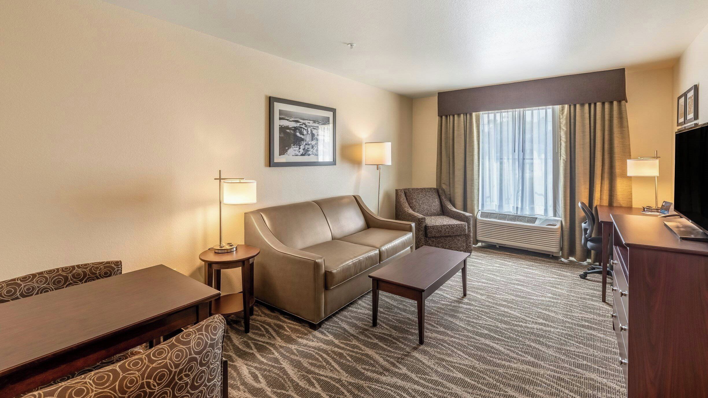 Ein Hotelzimmer mit Sofa, Ohrensessel, Schreibtisch und einem großen Fenster mit Vorhängen.