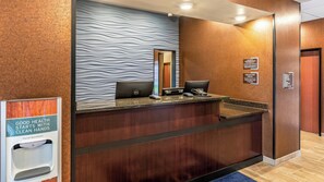 A hotel reception desk with a hand sanitizer dispenser, a mirror, and a sign indicating check-in and check-out times. - Cobblestone Hotel & Suites - Hartford (Hartford)