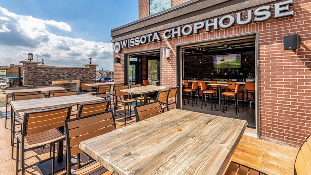 An outdoor seating area with wooden tables and chairs, a brick building with a sign reading 'WISSOTA CHOPHOUSE,' and a bar area visible through the entrance.