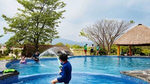 Una piscina con gente, una estructura de techo de paja y árboles al fondo.