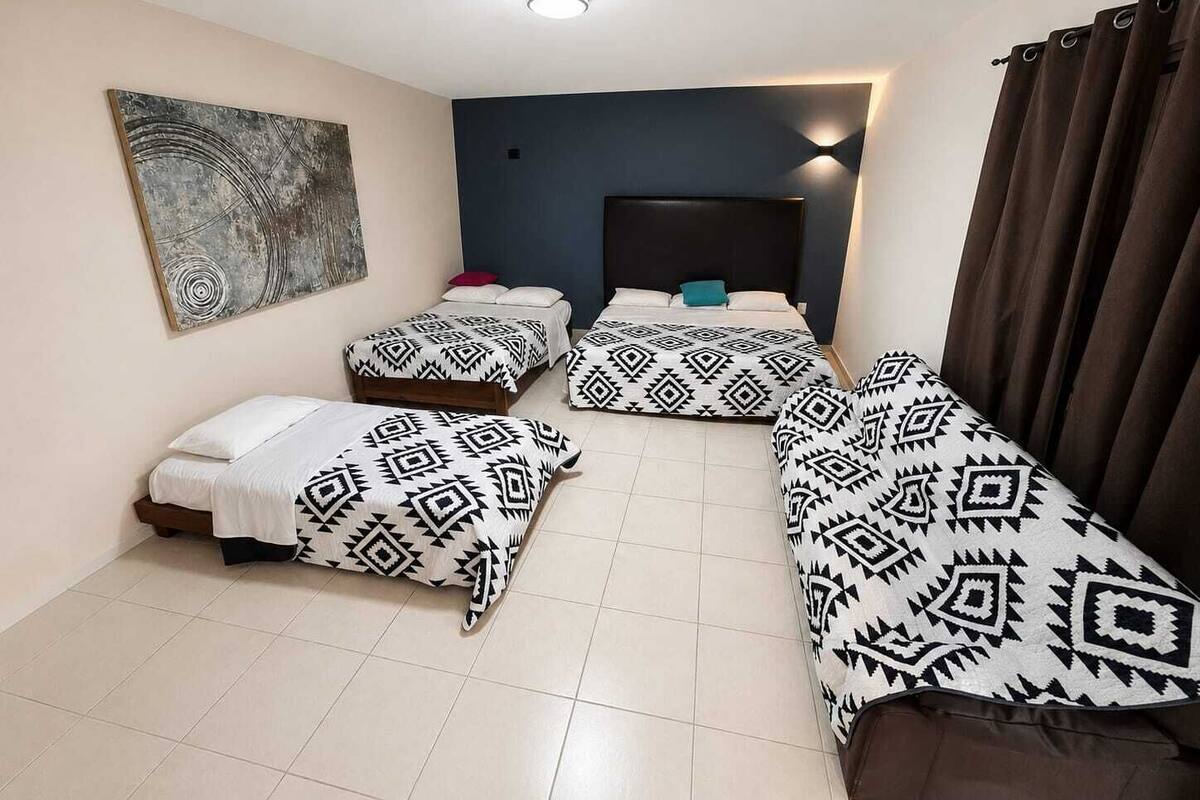 Een kamer met drie bedden, een donker hoofdbord, een aan de muur bevestigd abstract schilderij en een gordijn aan de rechterkant.