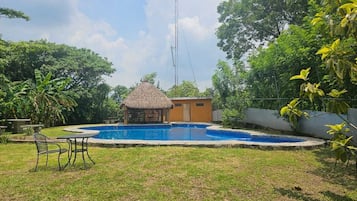 Una piscina con una cabaña de techo de paja, rodeada de follaje y un cielo despejado.