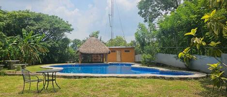 Una piscina con una cabaña de techo de paja, rodeada de follaje y un cielo despejado.
