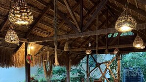 Een buitenterras met houten tafels en stoelen, rieten dak, hangende lampen en groen op de achtergrond.